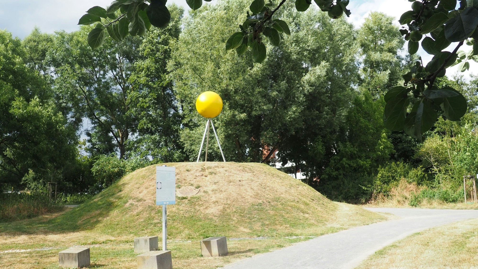 Auf einem kleinen Hügel, der nur spärlich mit Gras bewachsen ist, steht eine große gelbe Kugel auf drei Metallstangen.