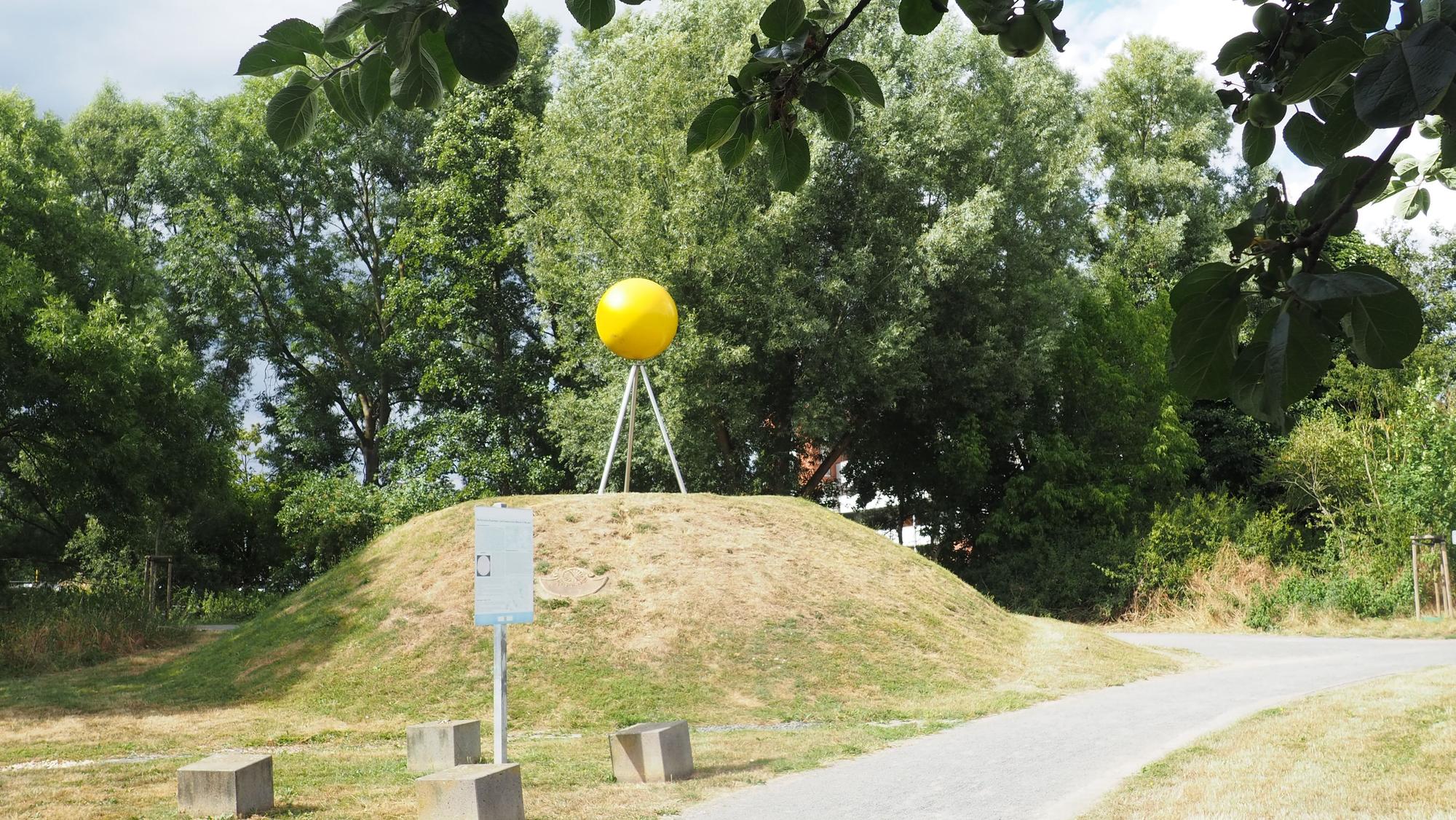 Auf einem kleinen Hügel, der nur spärlich mit Gras bewachsen ist, steht eine große gelbe Kugel auf drei Metallstangen.