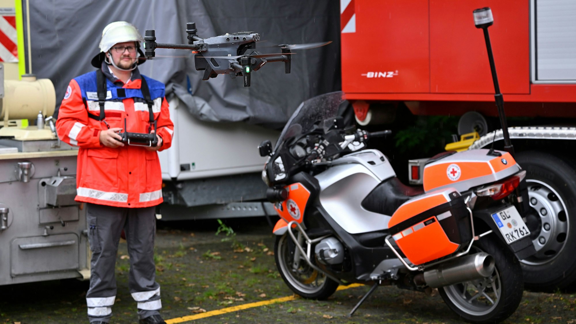 Ein DRK-Mann hat eine Fernbedienung in der Hand, eine Drohne hebt vom Boden ab. Neben dem Start- und Landeplatz steht ein DRK-Motorrad mit Blaulicht.