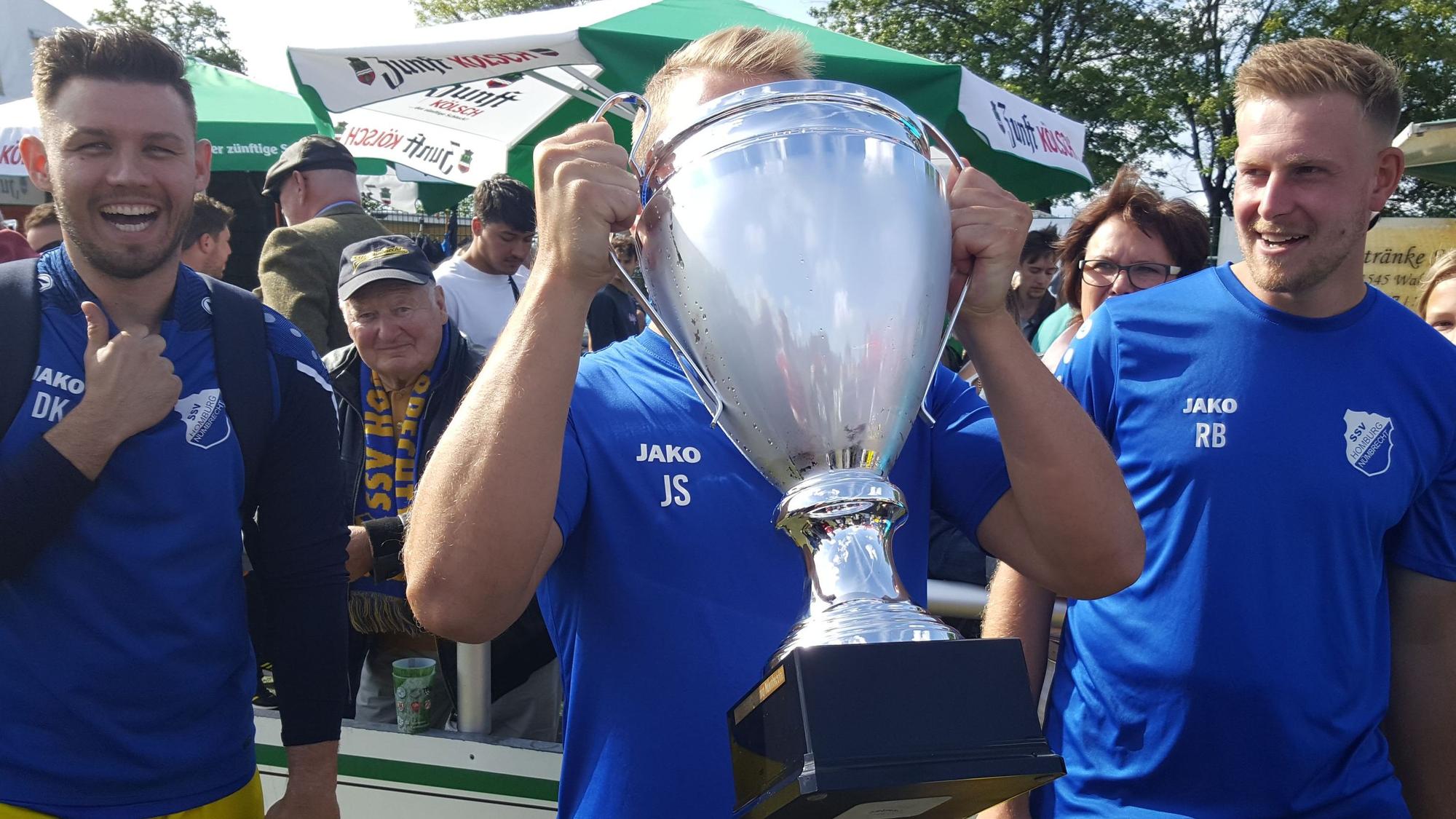 Drei Nümbrechter Fußballer freuen sich über den Sieg, in der Mitte nimmt einer einen tiefen Schluck aus dem Pokal.
