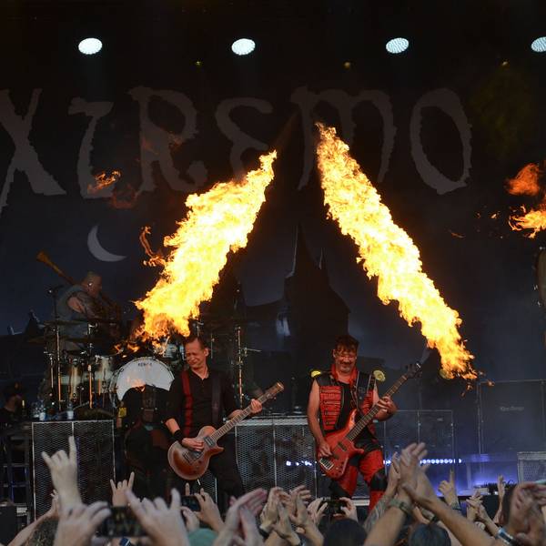 Eine Band steht auf einer Bühne, zur Show gehören auch Feuer-Fontänen. Im Vordergrund recken zahlreiche Fans die Hände in die Höhe.