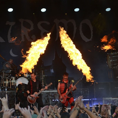 Eine Band steht auf einer Bühne, zur Show gehören auch Feuer-Fontänen. Im Vordergrund recken zahlreiche Fans die Hände in die Höhe.