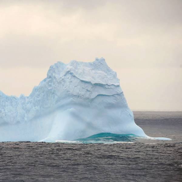 Ein Eisberg treibt im südlichen Ozean unweit der Antarktis im Ozean. Um ihn herum sind Wellen zu sehen, der Eisberg schimmert bläulich.