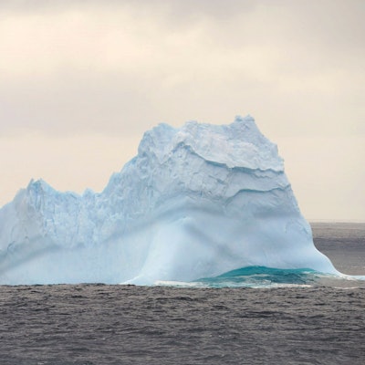 Ein Eisberg treibt im südlichen Ozean unweit der Antarktis im Ozean. Um ihn herum sind Wellen zu sehen, der Eisberg schimmert bläulich.