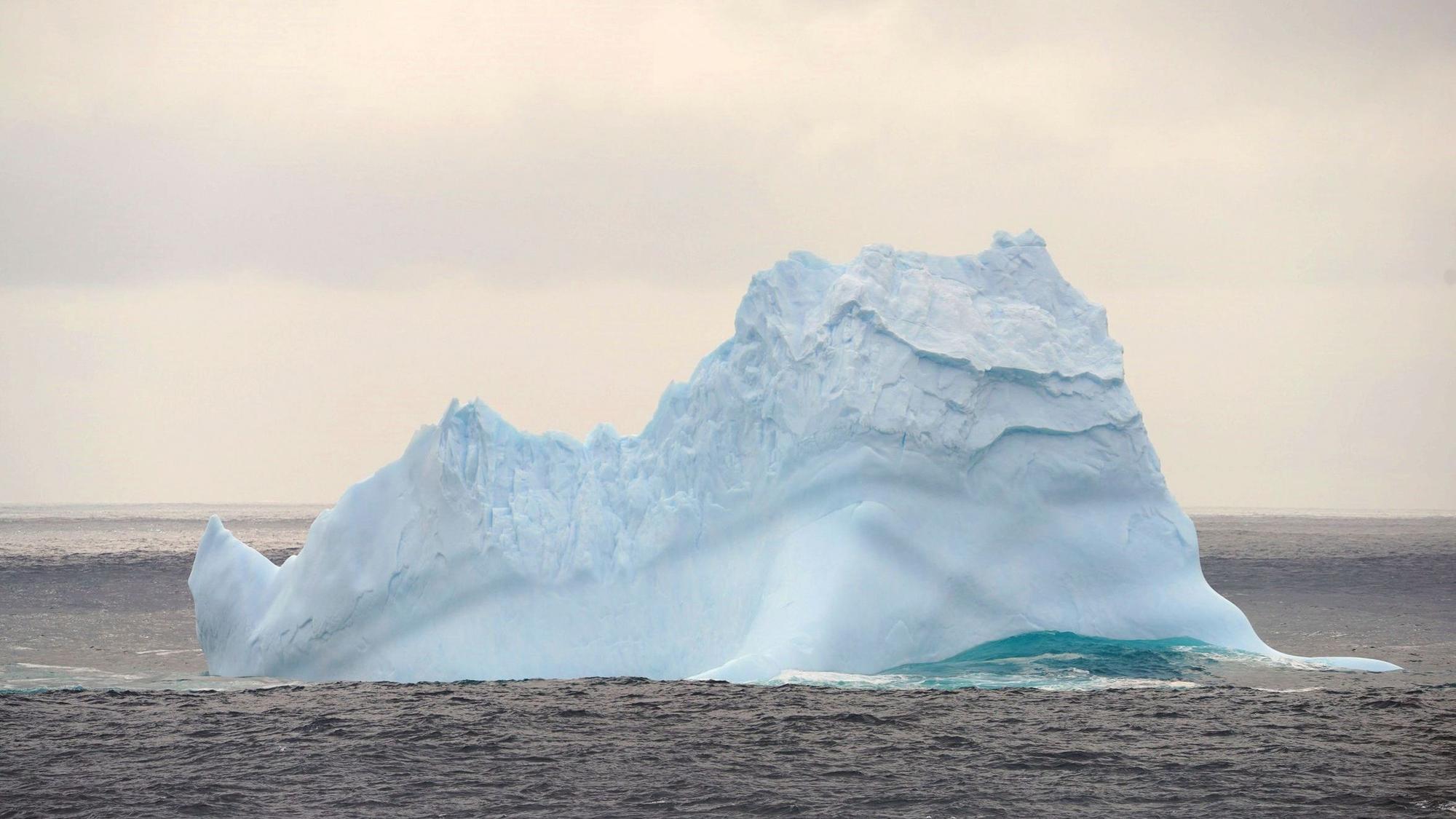 Ein Eisberg treibt im südlichen Ozean unweit der Antarktis im Ozean. Um ihn herum sind Wellen zu sehen, der Eisberg schimmert bläulich.