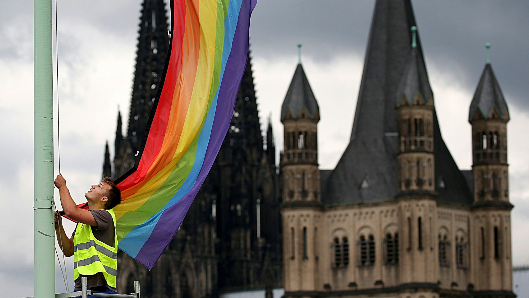ARCHIV - 30.06.2017, Nordrhein-Westfalen, Köln: Ein Arbeiter hängt nach der Entscheidung im Bundestag über die "Ehe für alle" vor der Kulisse des Doms und der Kirche Groß St. Martin (r) eine Regenbogenfahne auf.