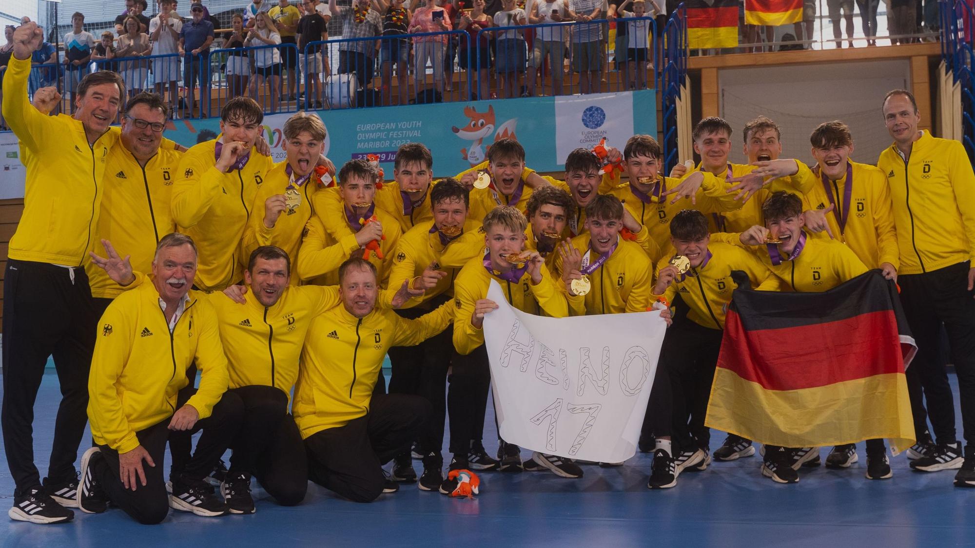 Die Handballer der deutschen U17 Nationalmannschaft jubeln nach dem Sieg beim EYOF mit ihren Goldmedaillen und denken mit einem Plakat an den verletzten Julius Hein.