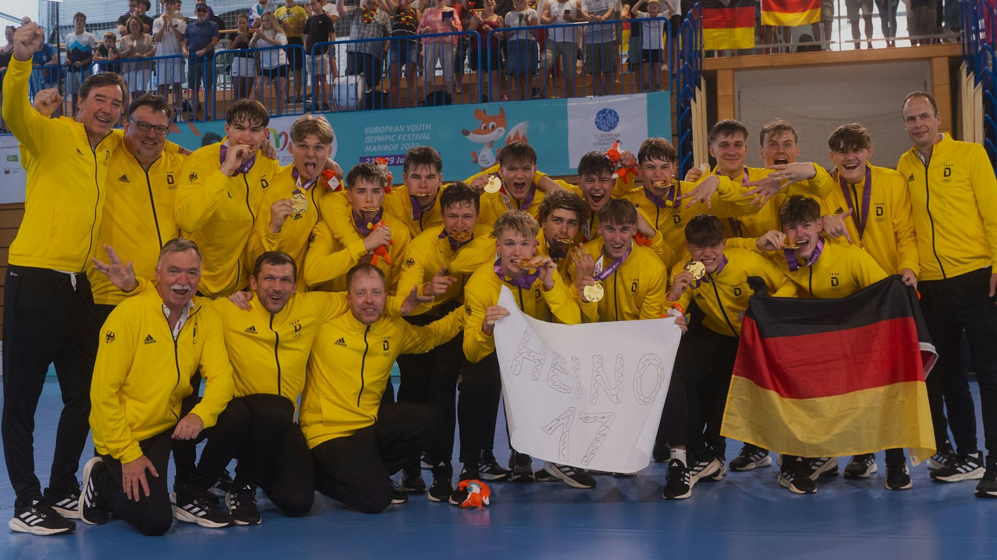 Die Handballer der deutschen U17 Nationalmannschaft jubeln nach dem Sieg beim EYOF mit ihren Goldmedaillen und denken mit einem Plakat an den verletzten Julius Hein.