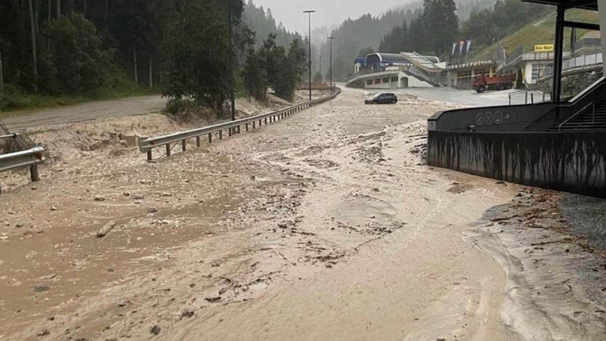 Schlamm und Wasser bedecken eine Straße in Südtirol. Im Hintergrund steckt ein Auto im Schlamm im Oberen Pustertal fest.