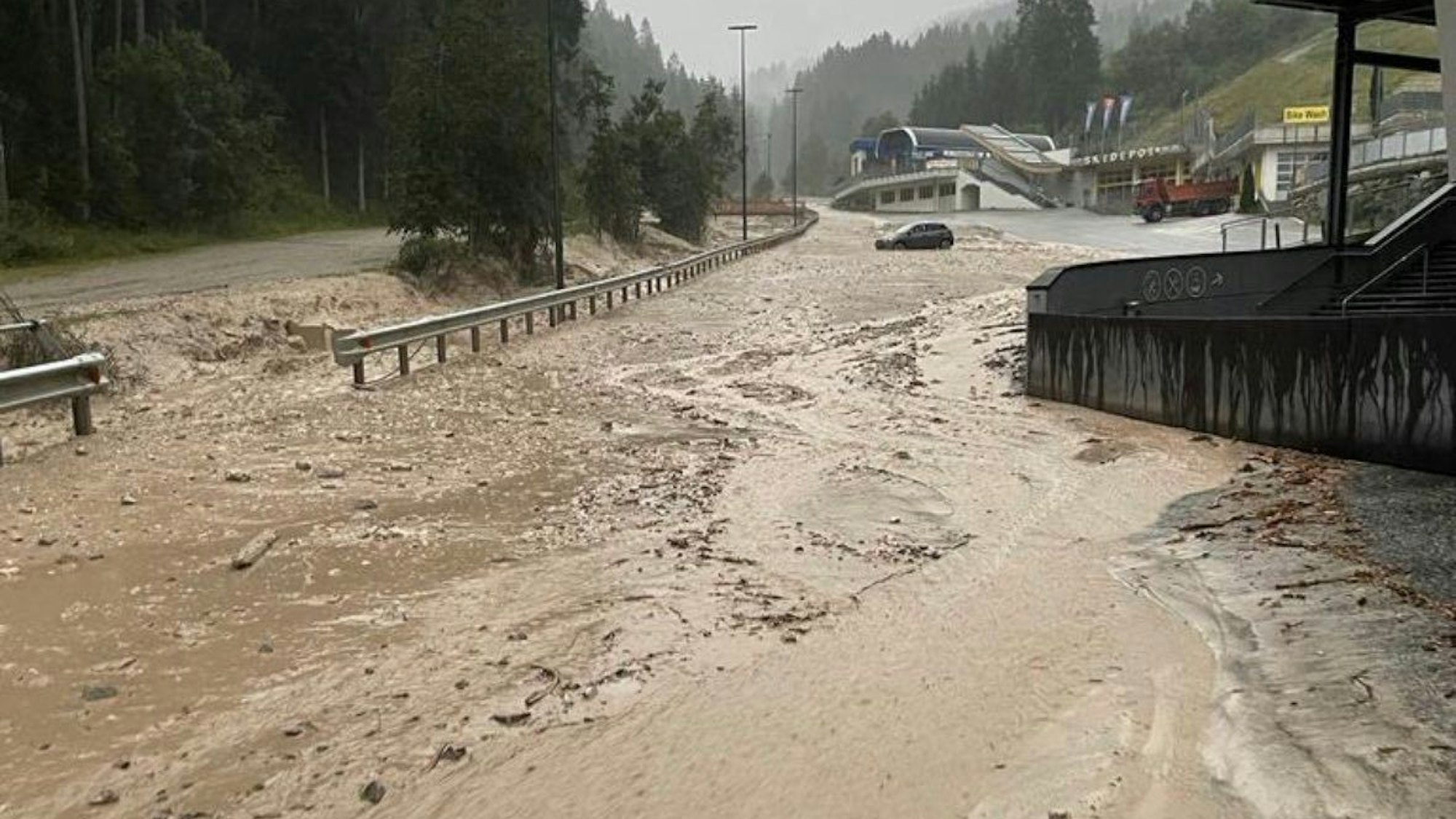 Schlamm und Wasser bedecken eine Straße in Südtirol. Im Hintergrund steckt ein Auto im Schlamm im Oberen Pustertal fest.