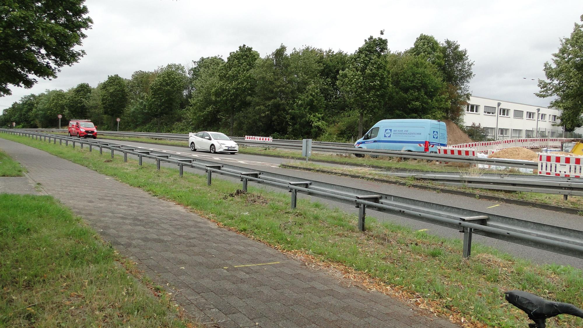 Autos fahren über eine Landstraße, ein Radweg ist durch Leitplanken abgetrennt.