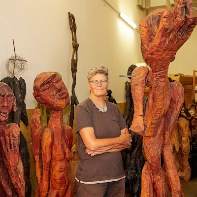 Die Künstlerin Rendel Freude steht inmitten ihrer Holzfiguren mit wulstigen Lippen und filigranen Gliedern im Figurenlager.