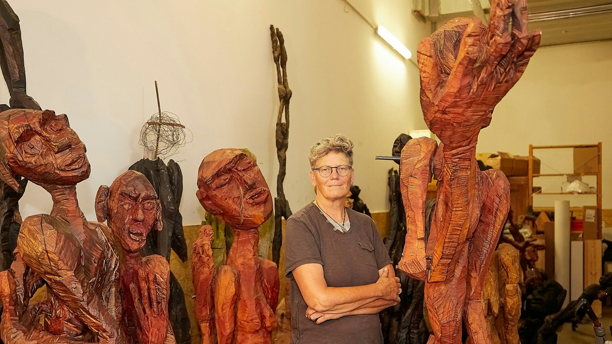 Die Künstlerin Rendel Freude steht inmitten ihrer Holzfiguren mit wulstigen Lippen und filigranen Gliedern im Figurenlager.
