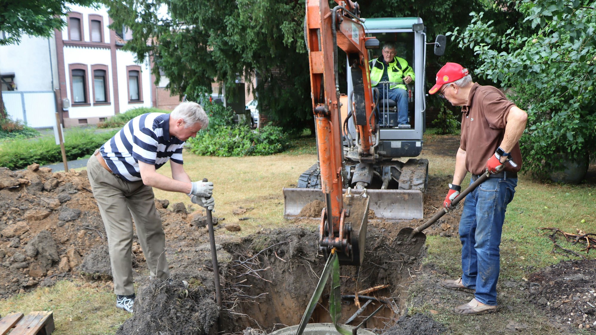 Ein Mann auf einem Bagger in der Mitte und zwei Männer mit Schaufeln links und rechts daneben heben ein Loch aus.