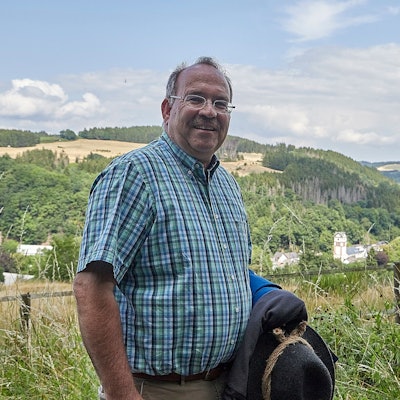 Ein Mann im rustikalen Wanderoutfit steht vor einer sommerlichen Eifellandschaft.