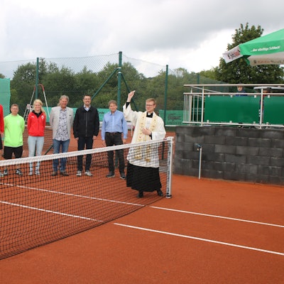 Mehrere Menschen stehen auf einem Tennisplatz. Ein Kaplan weiht den Platz mit Weihwasser.