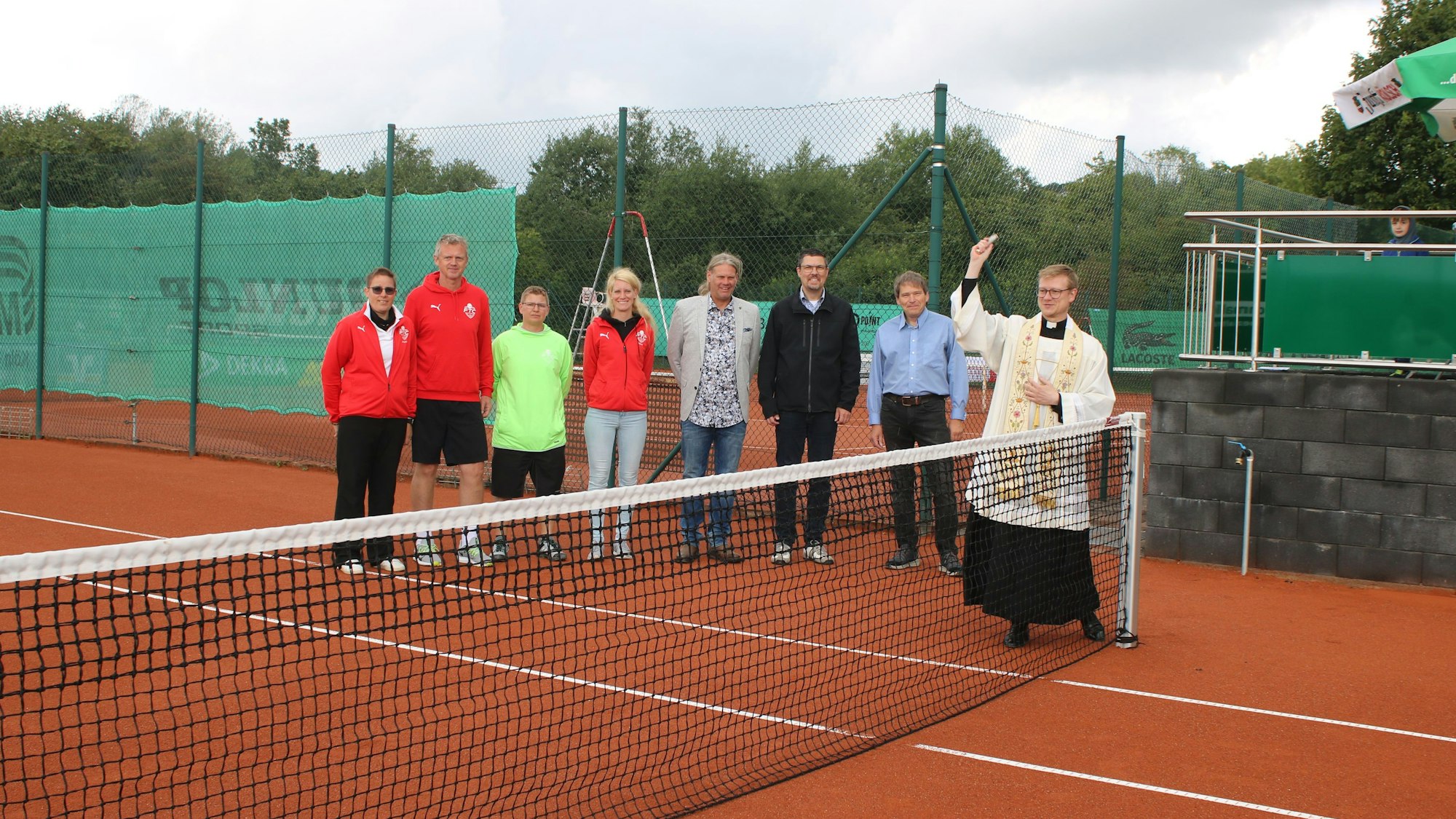 Mehrere Menschen stehen auf einem Tennisplatz. Ein Kaplan weiht den Platz mit Weihwasser.
