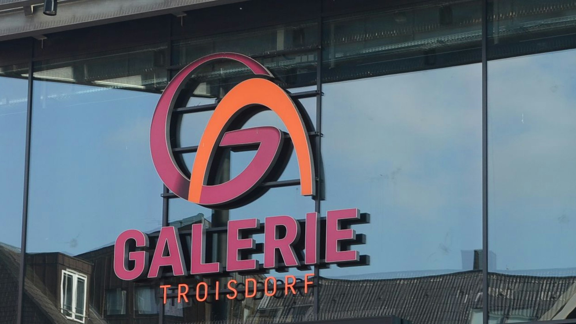 Leuchtreklame für die Galerie Troisdorf in Weinrot und Orange; in der Glasscheibe spiegeln sich Dächer.