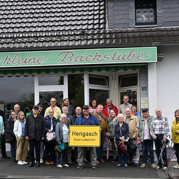 Menschen stehen mit einer gelben Ortstafel, auf der zu lesen ist „Hengasch Kreis Liebernich“ vor einem Ladenlokal mit der Aufschrift „Die kleine Backstube“.