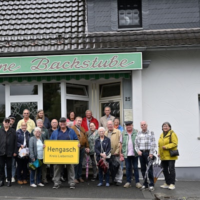 Menschen stehen mit einer gelben Ortstafel, auf der zu lesen ist „Hengasch Kreis Liebernich“ vor einem Ladenlokal mit der Aufschrift „Die kleine Backstube“.