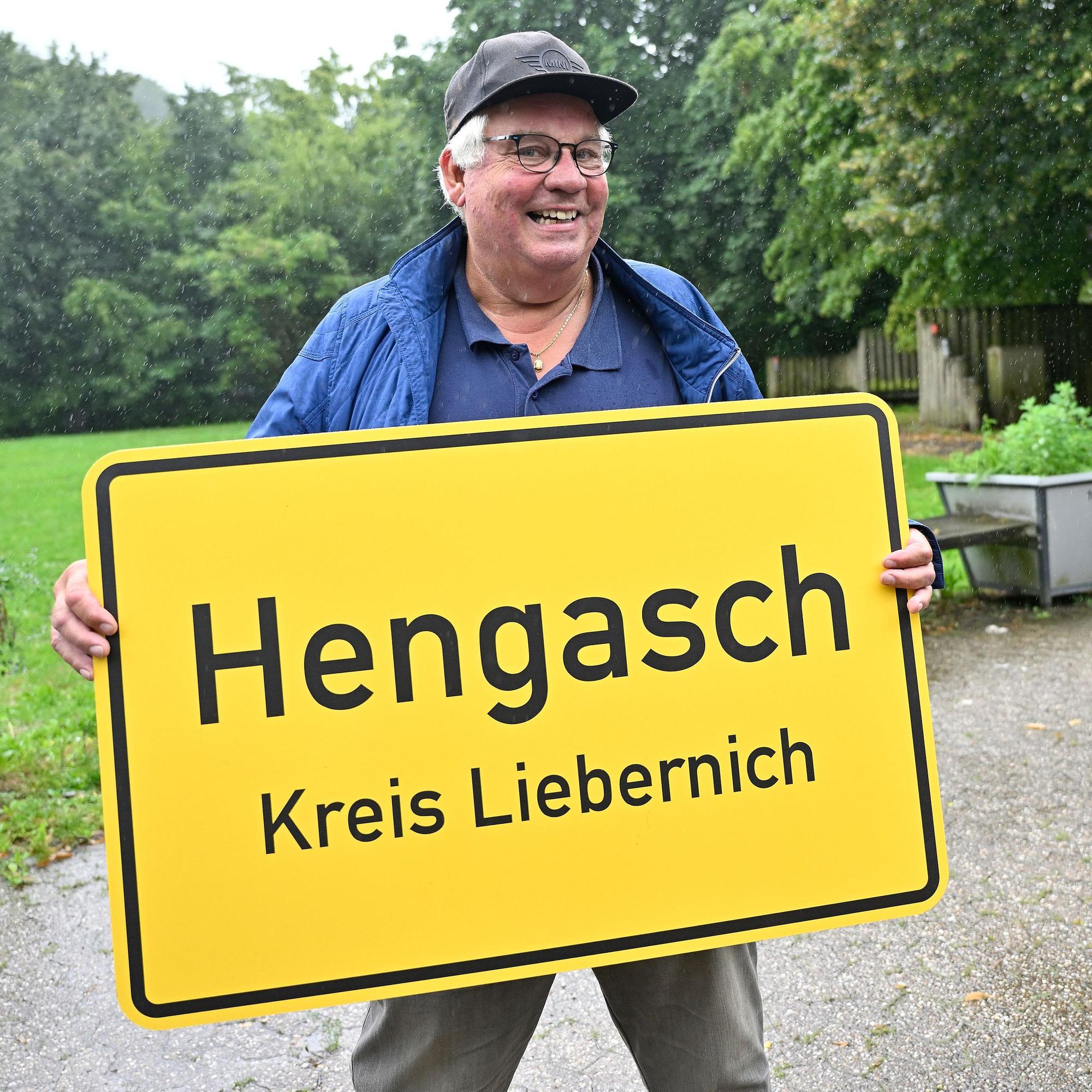 Ein Mann hält eine gelbe Ortstafel in Händen, auf dieser steht: Hengasch Kreis Liebernich.