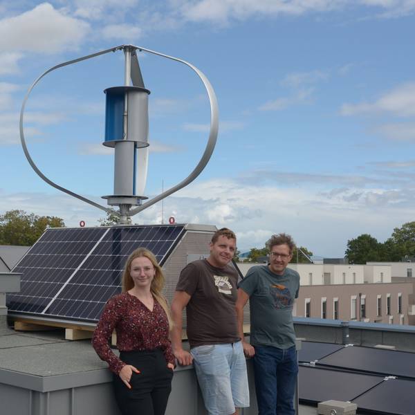 Zwei Männer und eine Frau stehen auf dem Dach eines Hauses vor einem Wind-Solar-Kleinkraftwerk.