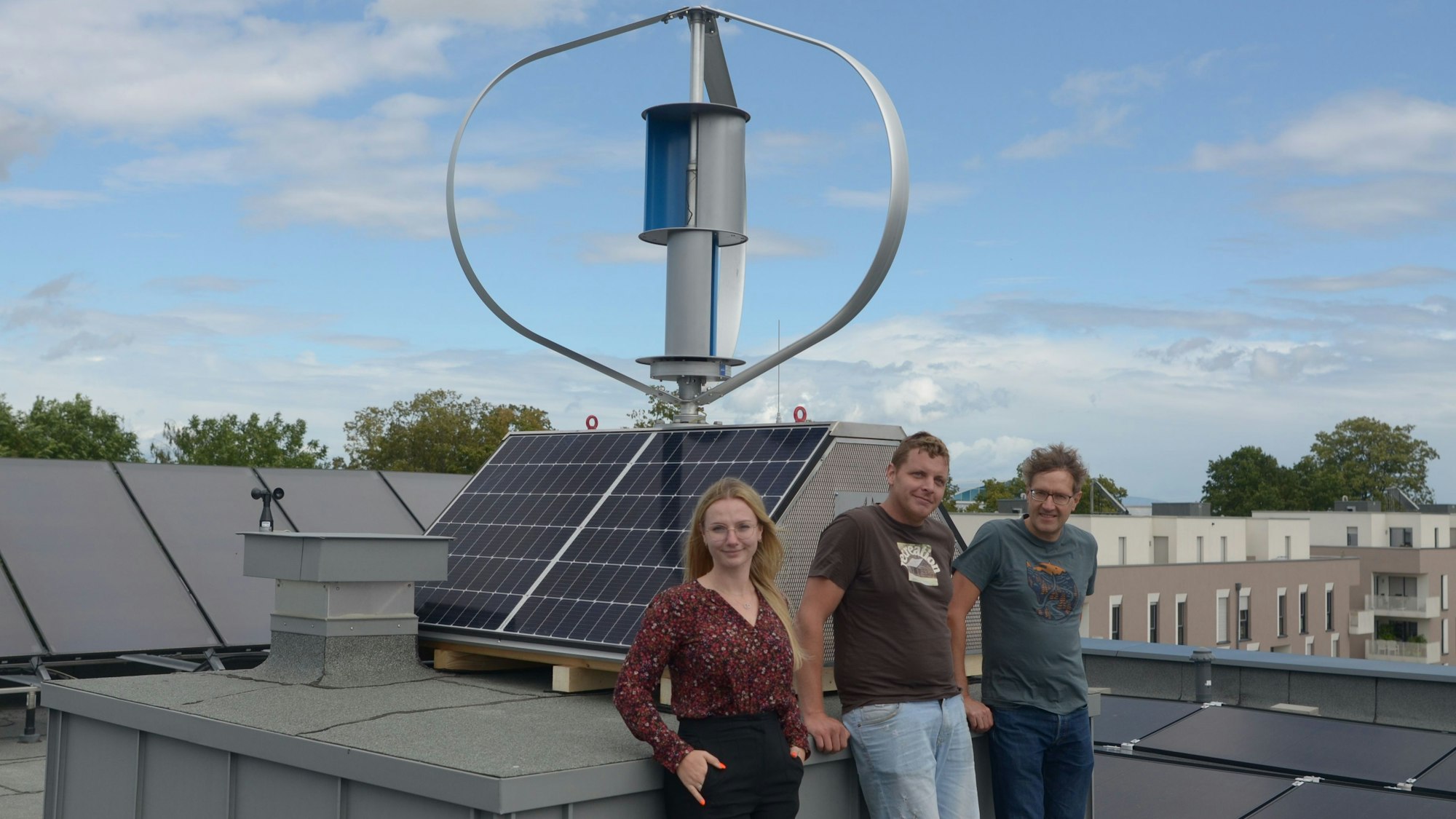 Zwei Männer und eine Frau stehen auf dem Dach eines Hauses vor einem Wind-Solar-Kleinkraftwerk.