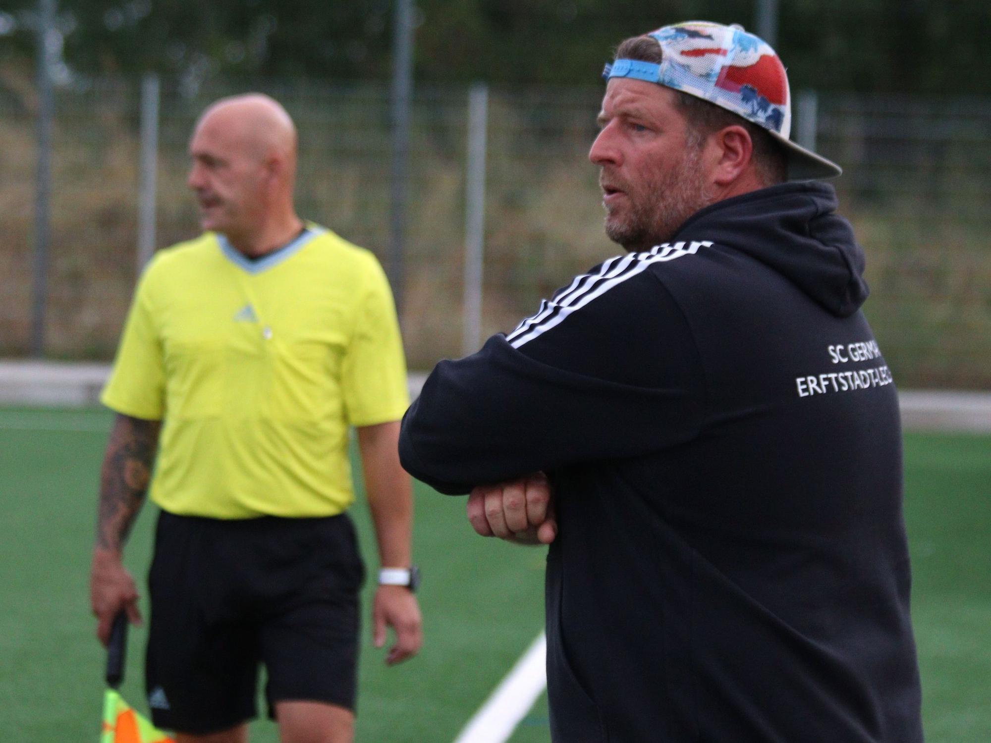 Karsten Kochems, Trainer von Erftstadt-Lechenich, trägt eine bunte Kappe verkehrt herum auf dem Kopf. Im Hintergrund Linienrichter Jörg Piana.