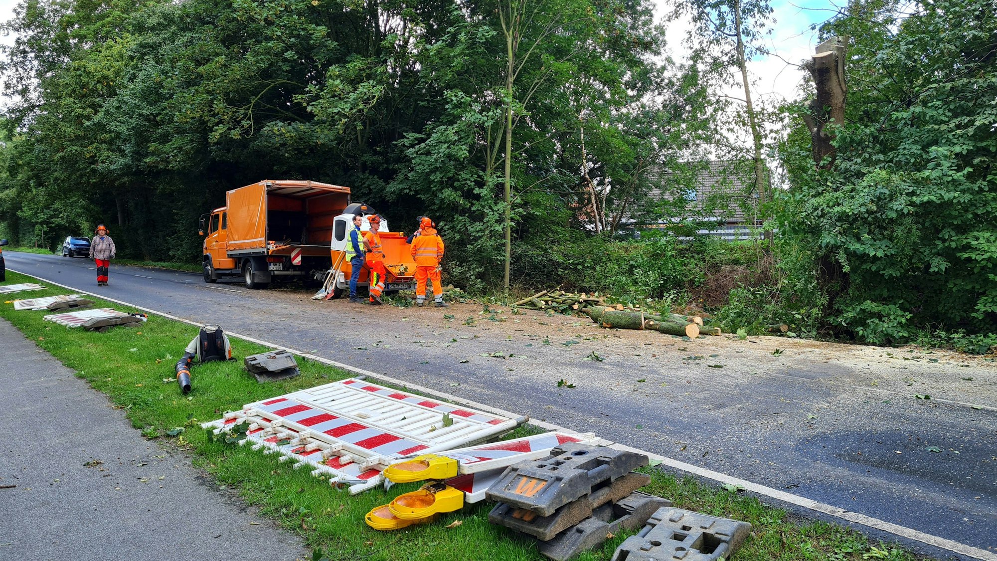 Im Vordergrund liegen rot-weiße Baken und andere Absperrelemente auf dem Grünstreifen neben einer Straße, im Hintergrund stehen Arbeiter auf der Straße neben einem Baumfahrzeug und einem gefällten Baum.