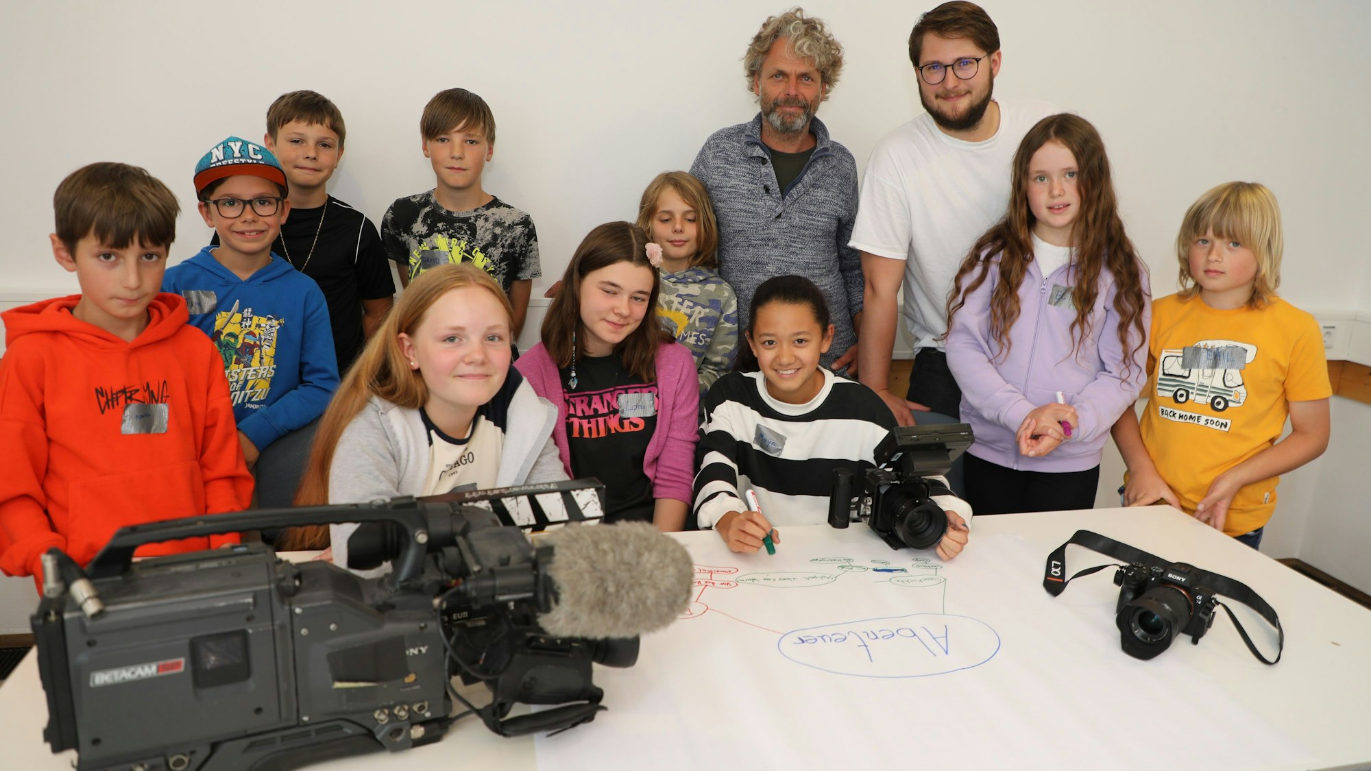 Kinder stehen mit zwei Erwachsenen hinter Filmequipment und sammeln Ideen auf einem großen Plakat.