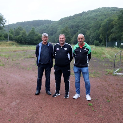 Die Verantwortlichen der SG Houverath/Mutscheid stehen auf dem Aschenplatz, der seit der Flut nicht mehr nutzbar ist und mittlerweile von Unkraut übersät ist. Neben ihnen liegt ein Kleinfeldtor.