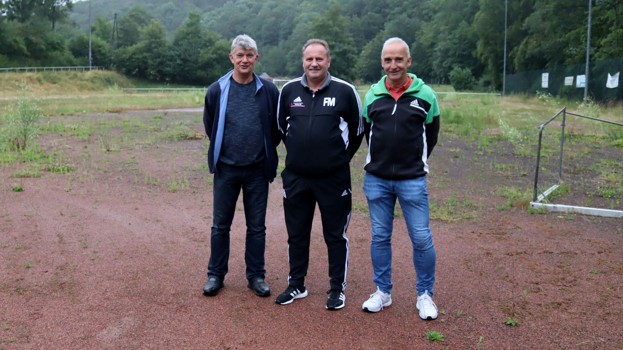 Die Verantwortlichen der SG Houverath/Mutscheid stehen auf dem Aschenplatz, der seit der Flut nicht mehr nutzbar ist und mittlerweile von Unkraut übersät ist. Neben ihnen liegt ein Kleinfeldtor.