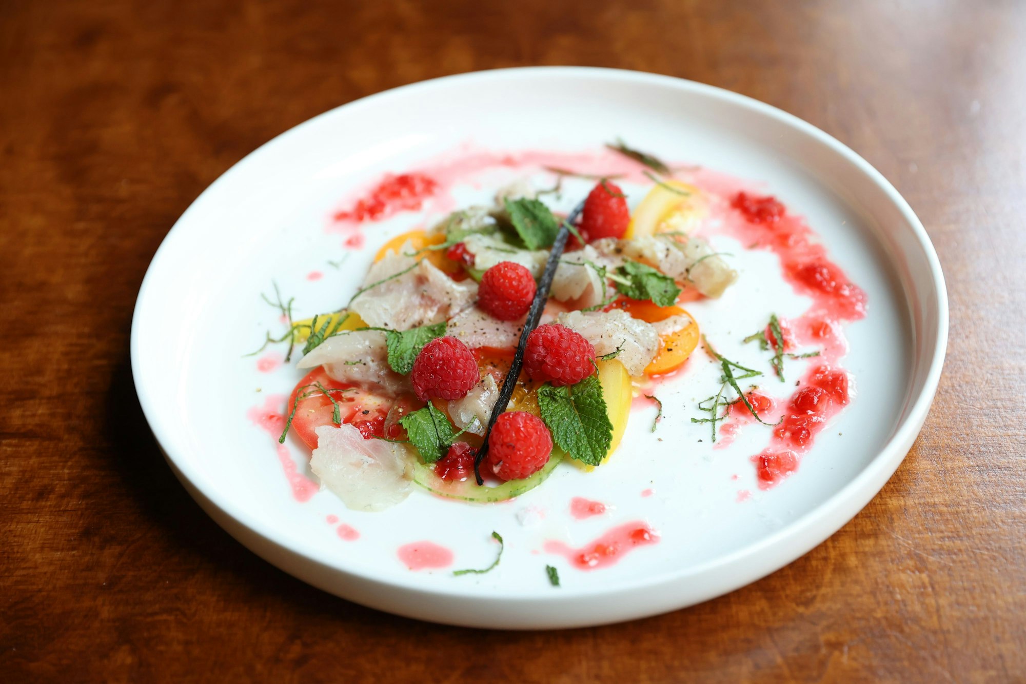 Roh marinierter Wolfsbarsch liegt garniert mit Vanille, bretonischen Tomaten, Himbeeren und Zitronenmelisse auf einem Teller.