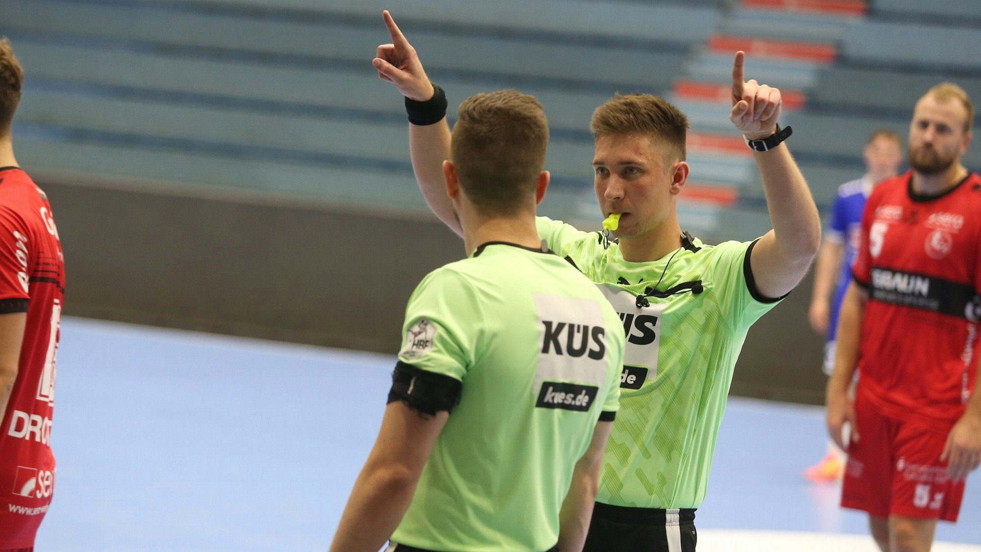 Zwei Schiedsrichter stehen auf einem Handballfeld, einer zeigt den Videobeweis an.