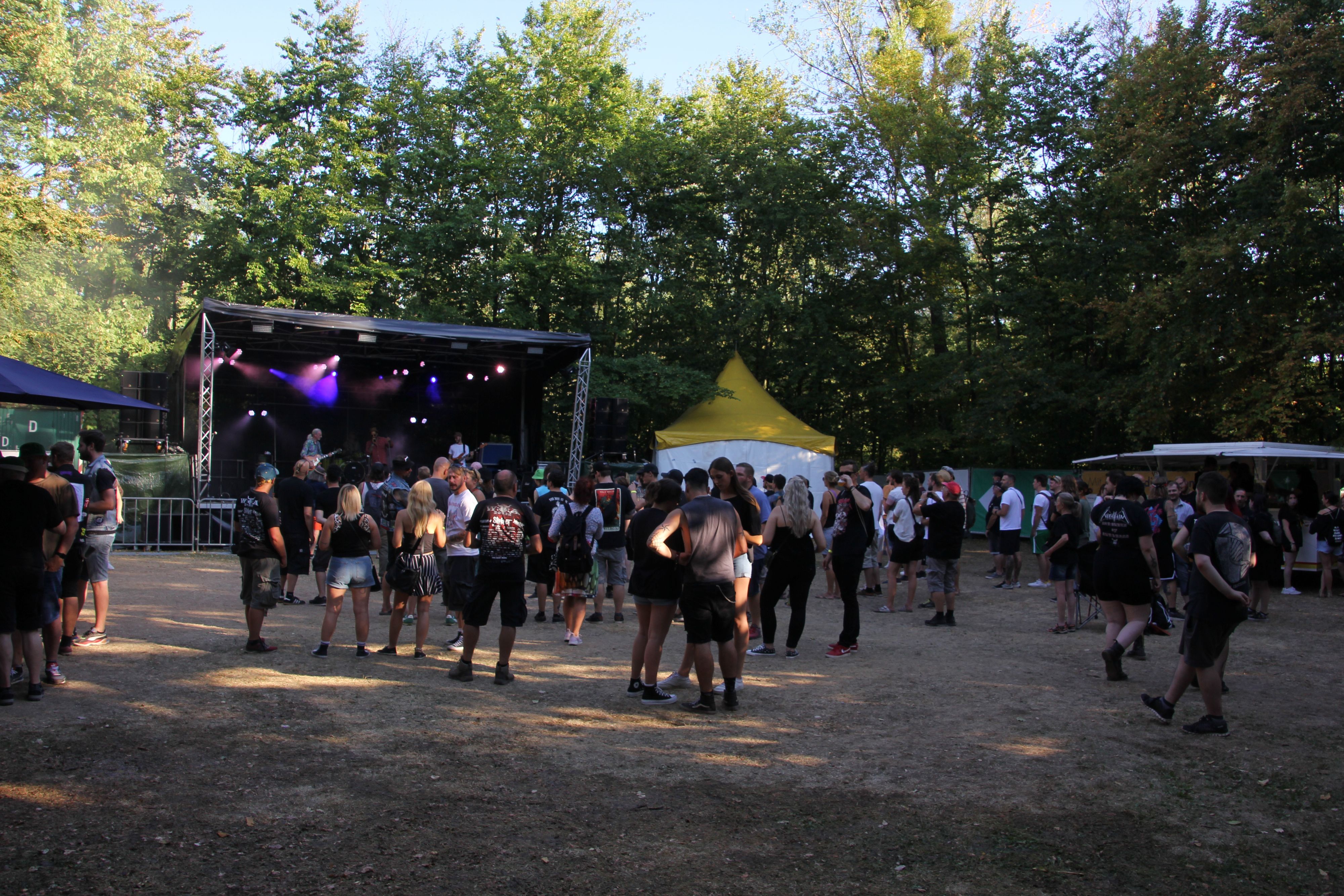 Vor einer kleinen Bühne im Wald stehen Fans und hören der Band zu.