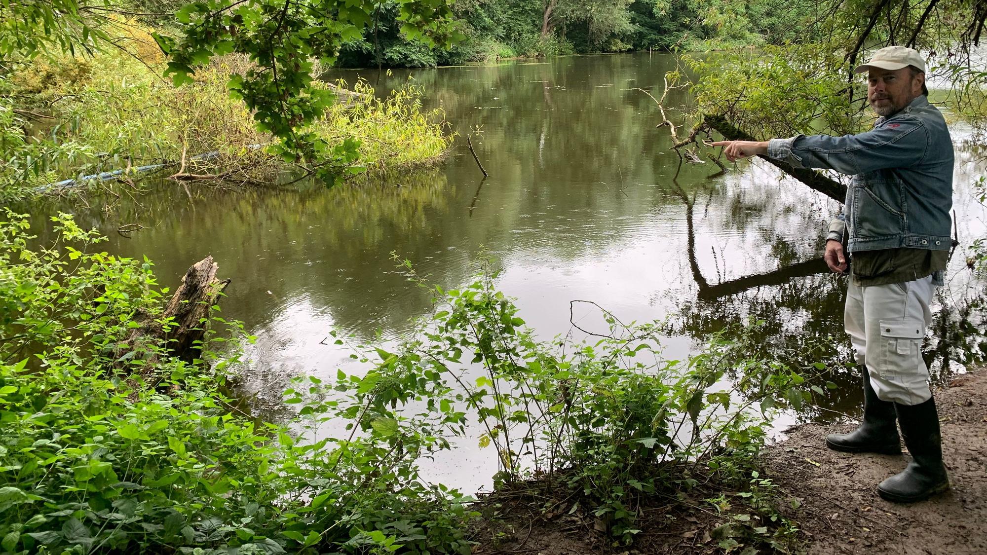 Ein Mann steht am Ufer eines Sees. Er deutet mit dem Finger auf das Wasser.