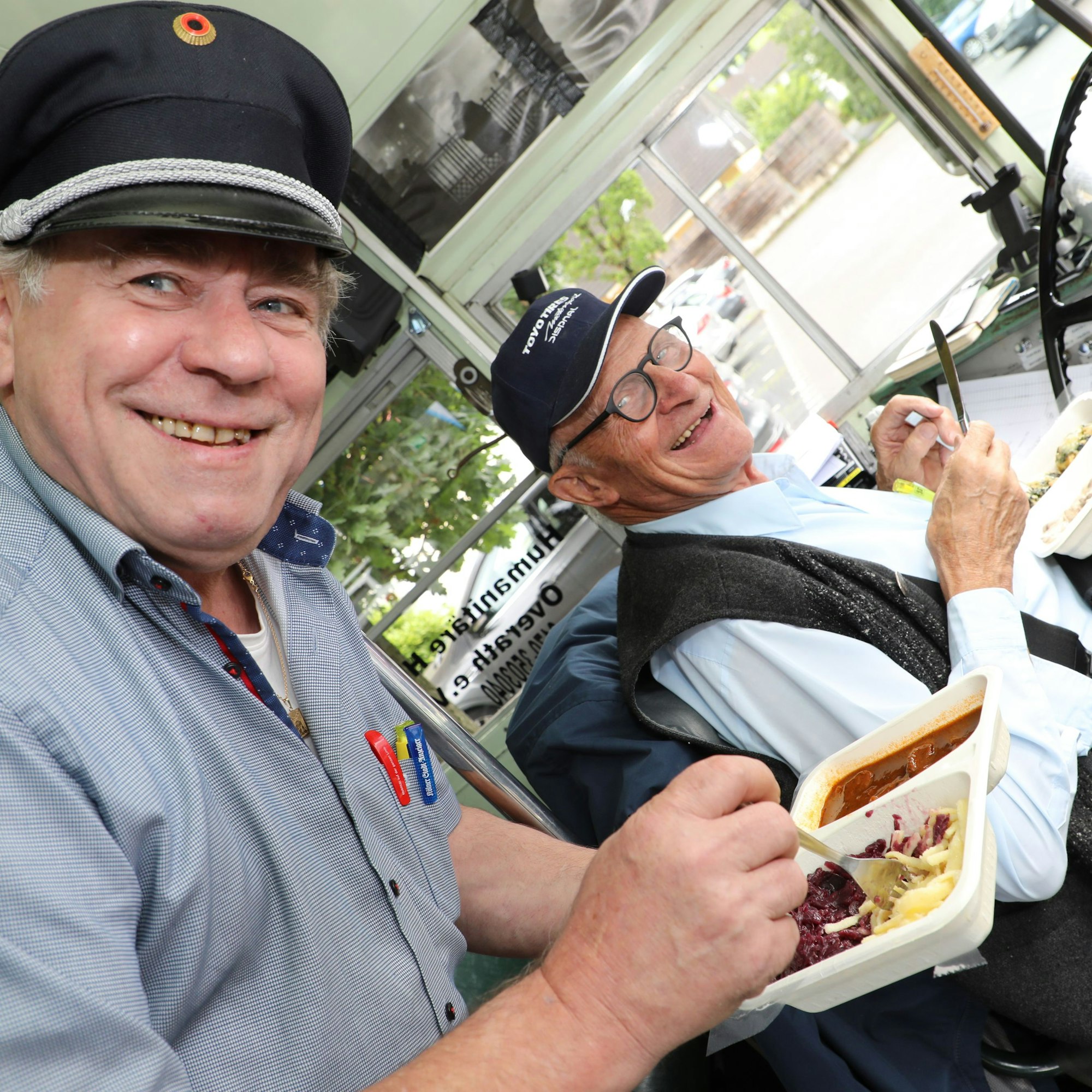 Zwei Männer im Cockpit eines historischen Busses essen ein Mittagsgericht aus Pappschalen.