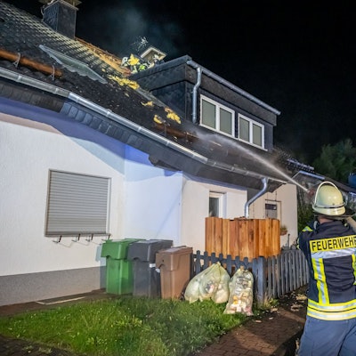 Ein Feuerwehrmann hält einen Schlauch, aus dem ein Wasserstrahl kommt, in Richtung eines brennenden Dachstuhls.
