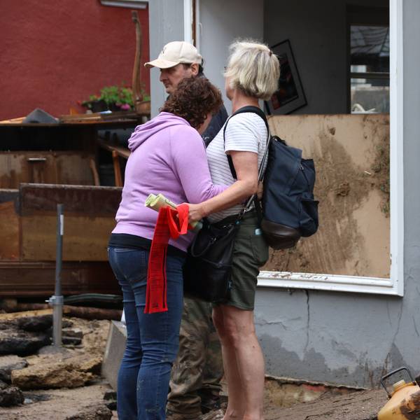 Das Bild zeigt zwei Frauen, die sich nach der Flutkatastrophe in Bad Münstereifel trösten. Im Hintergrund sind ein Mann und ein zerstörtes Haus zu sehen.