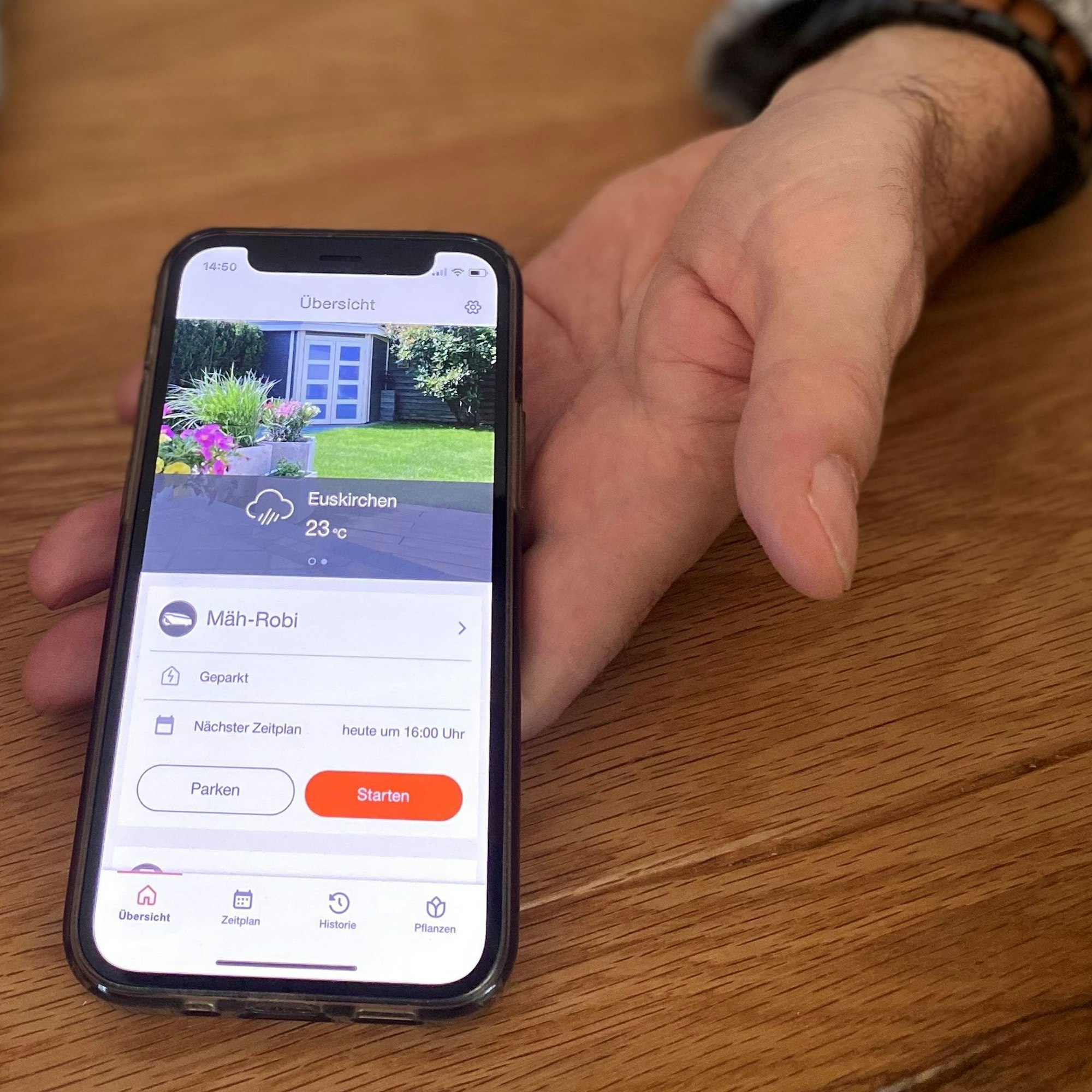 Eine Hand hält ein Smartphone. Auf dem Display ein Foto des Gartens der Euskirchener, darunter eine Temperaturanzeige und ein Regensymbol.