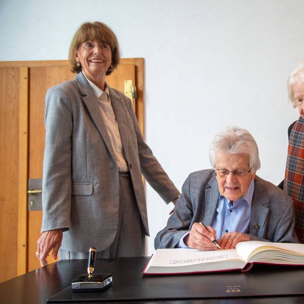 Peter Busmann, Vreneli Busmann und Henriette Reker stehen an einem Tisch vor dem Gästebuch der Stadt.