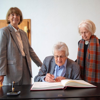 Peter Busmann, Vreneli Busmann und Henriette Reker stehen an einem Tisch vor dem Gästebuch der Stadt.