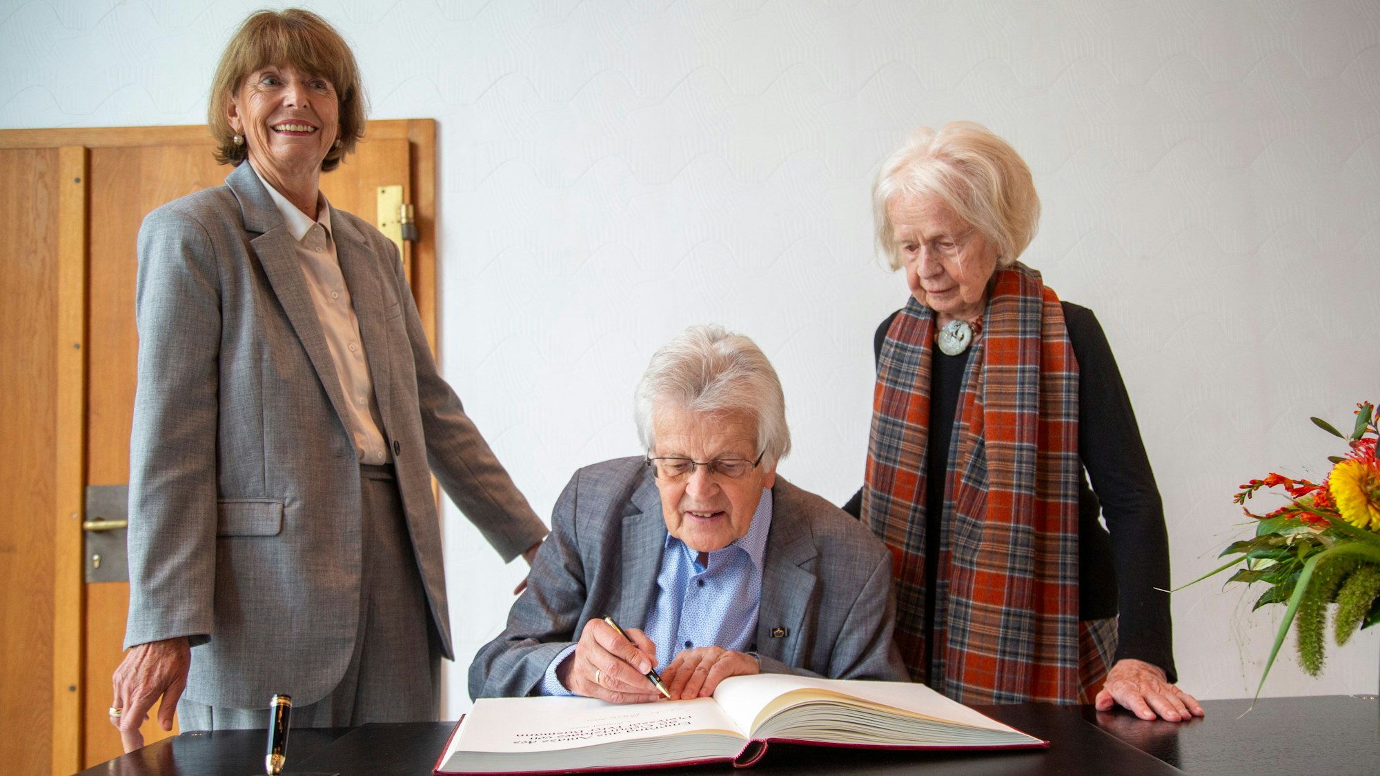 Peter Busmann, Vreneli Busmann und Henriette Reker stehen an einem Tisch vor dem Gästebuch der Stadt.