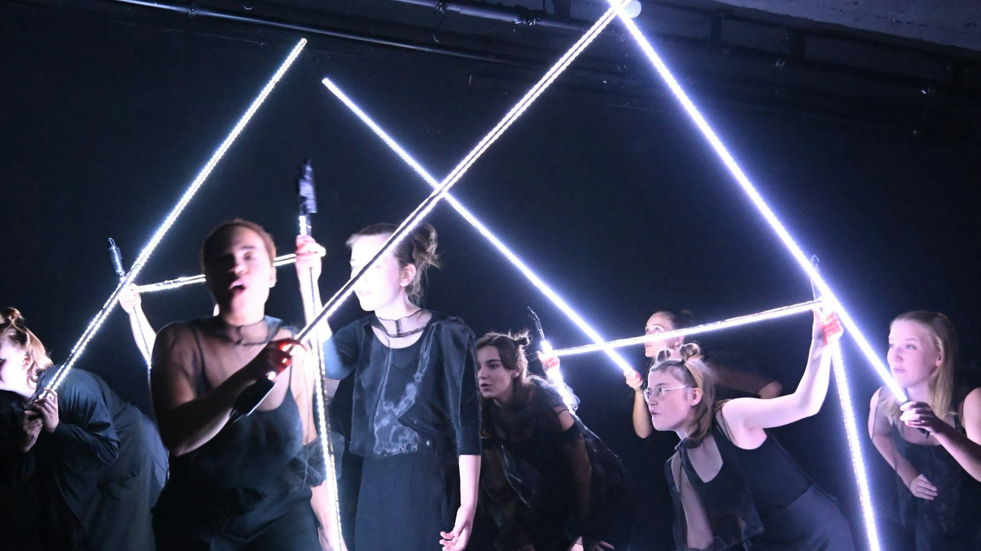 Jugendliche stehen auf der Bühne und halten sich an leuchtenden Stäben, die wie ein Haus aufgebaut sind, fest.