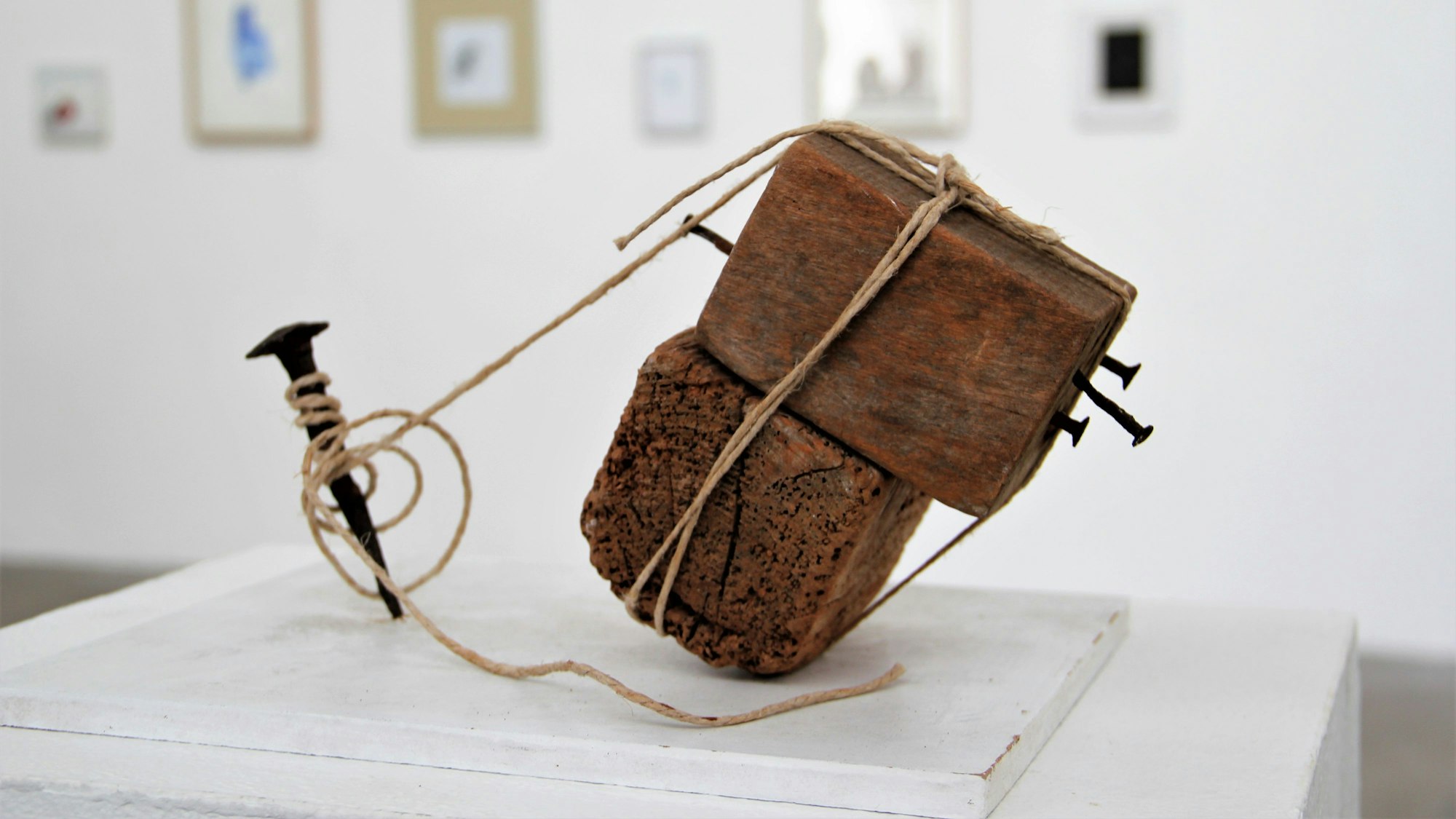 Ein Holzobjekt, das mit Bindfäden an einem Nagel befestigt ist, steht in einem Ausstellungsraum.