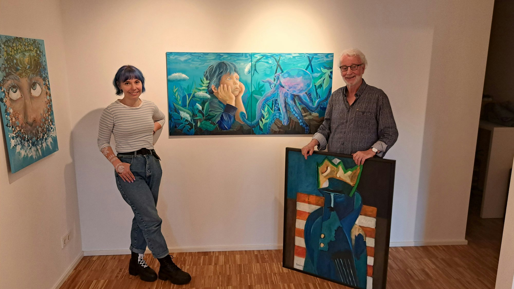 Ein Mann und eine Frau stehen in einer Galerie vor einem Bild. Der Mann hält ein weiteres Bild in den Händen.