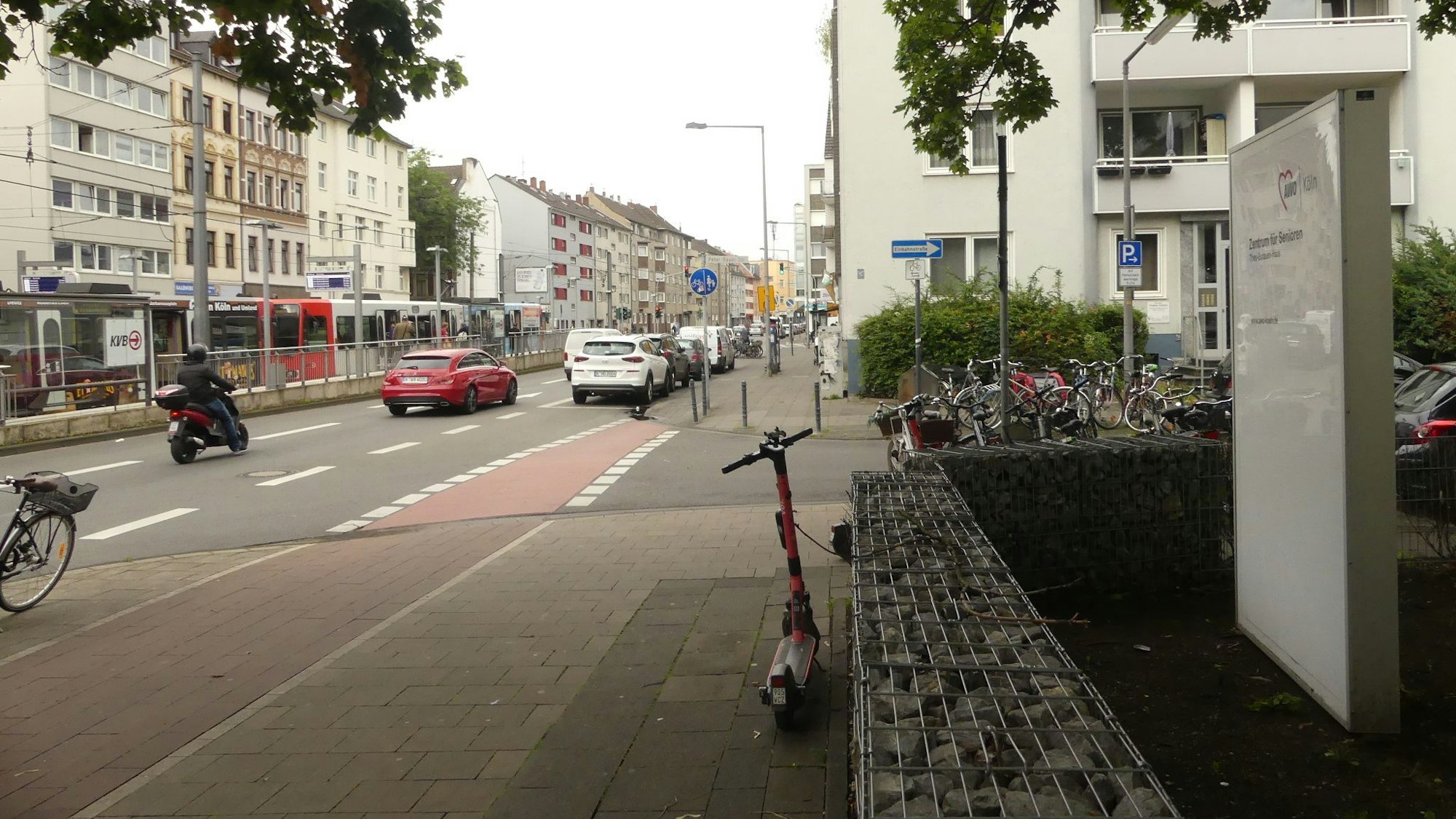 Eine Straße mit Auto-, Moped- und Straßenbahnverkehr ist zu sehen. Im Vordergrund seitlich steht das Schild des Theo-Burauen-Hauses, eines Altenheims.