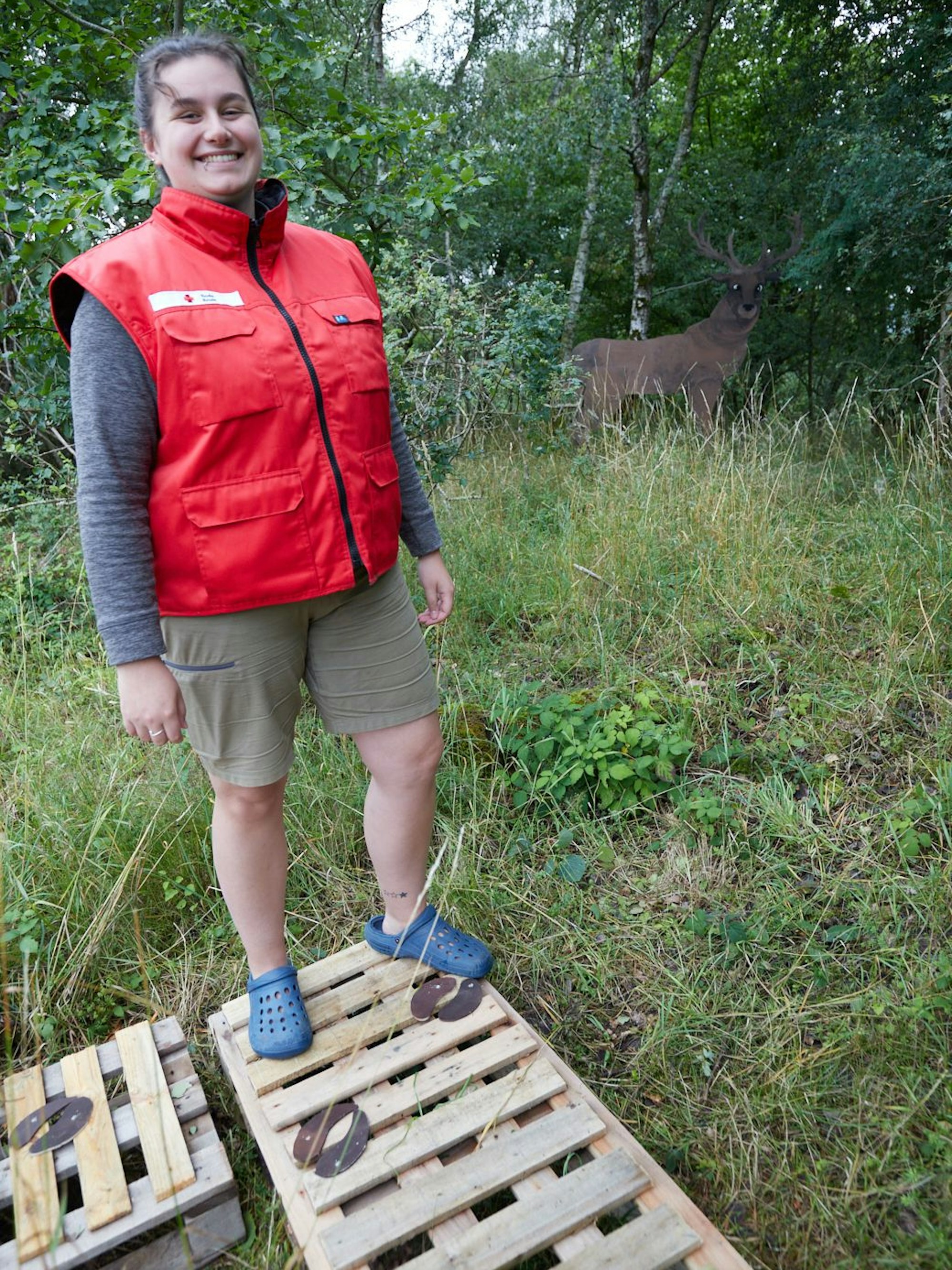Laetitia Lannois steht auf Paletten, die als Steg dienen. Im Hintergrund ist im Dickicht ein hölzerner Rothirsch zu sehen.