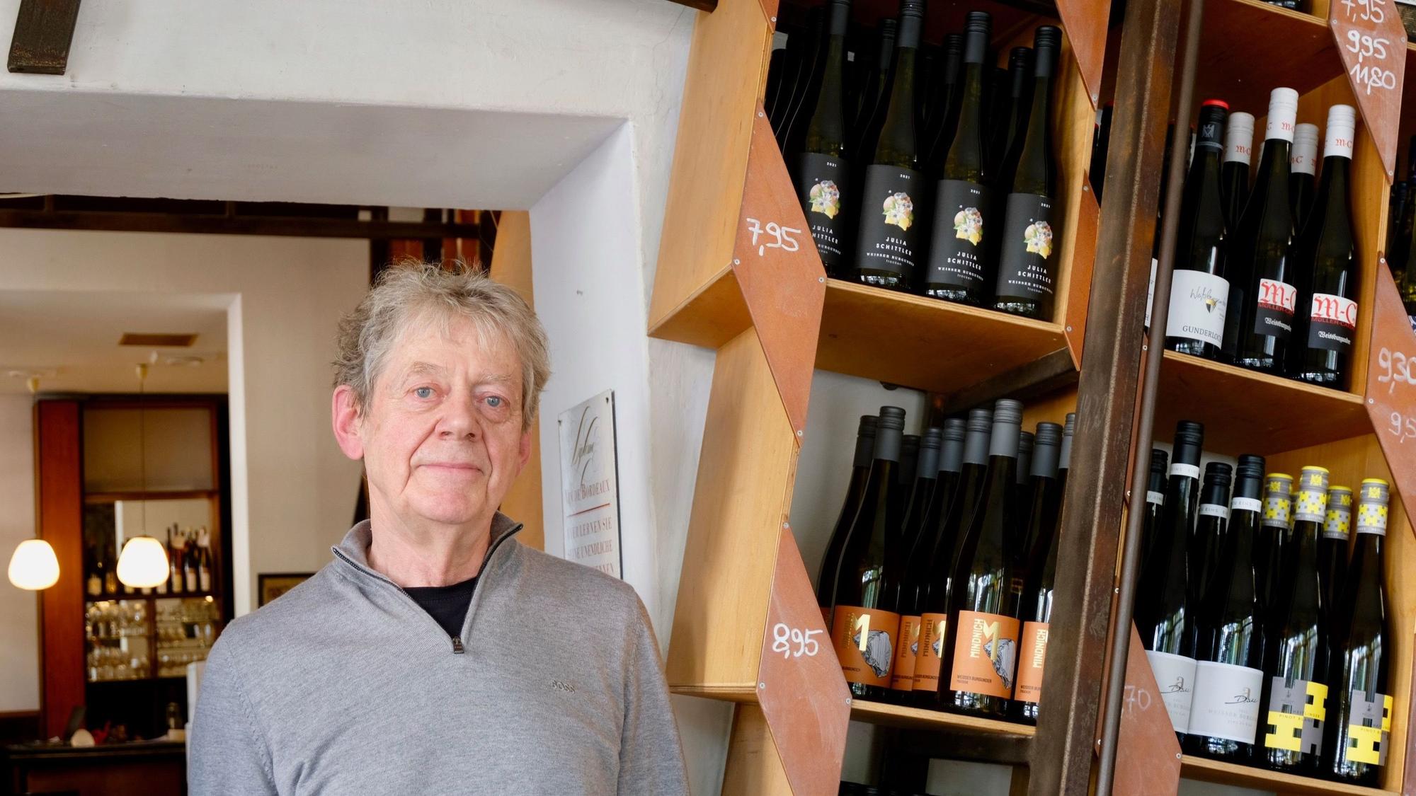Ein älterer Mann steht in einem Weinladen, neben ihm sind Regale mit Weinflaschen zu sehen.