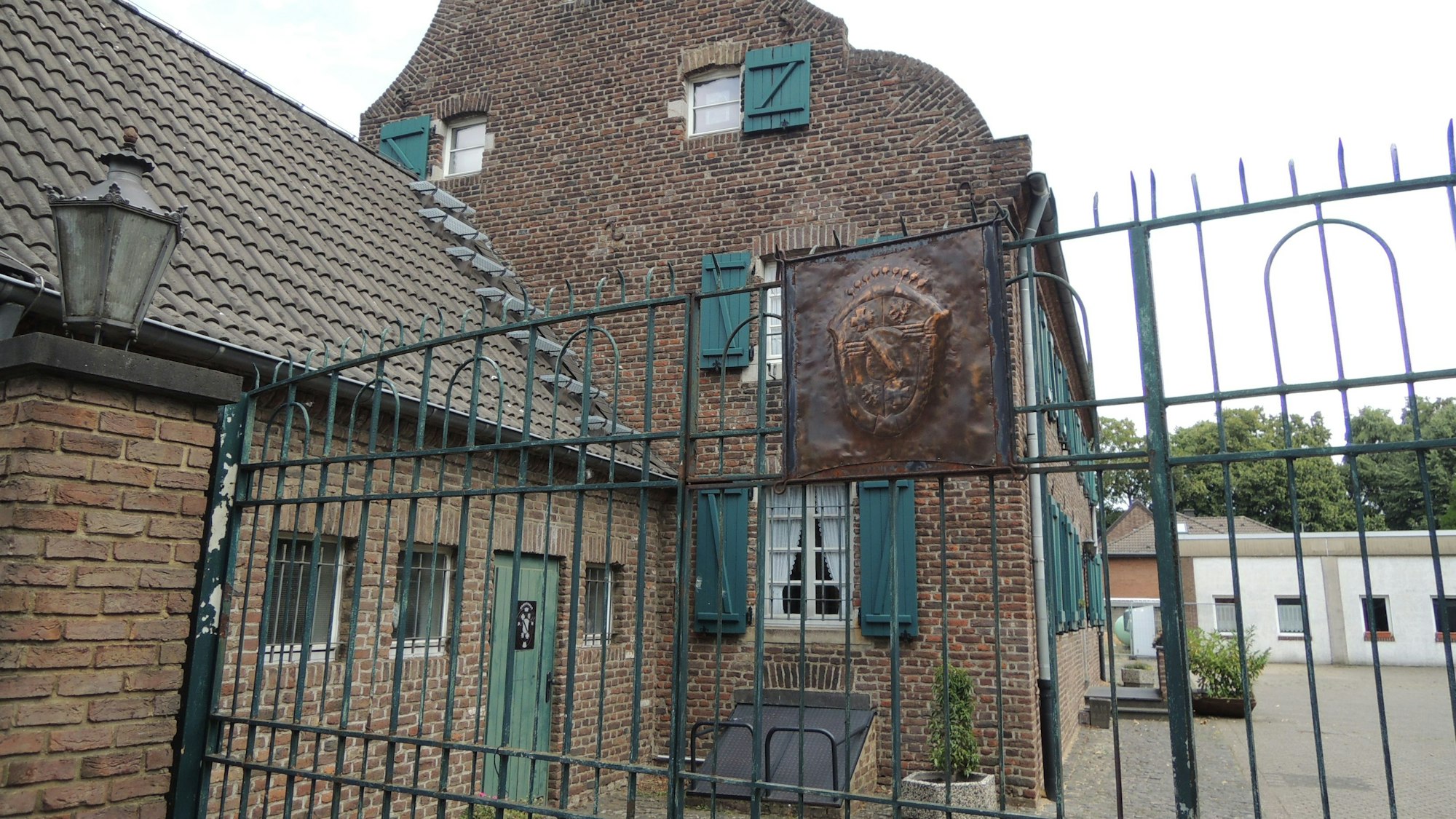 Hinter einem grünen Metalltor steht ein Backstein-Gebäude mit grünen Fensterläden.